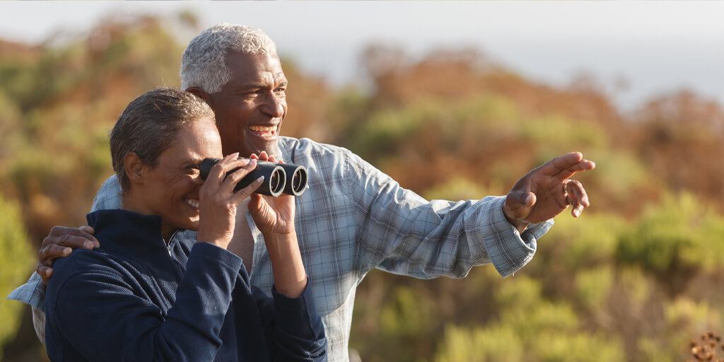 A senior couple outside pointing in the distance and looking through binoculars.