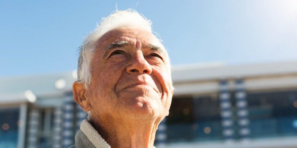 A senior man enjoys his retirement, standing outdoors and smiling up at the sunny sky.