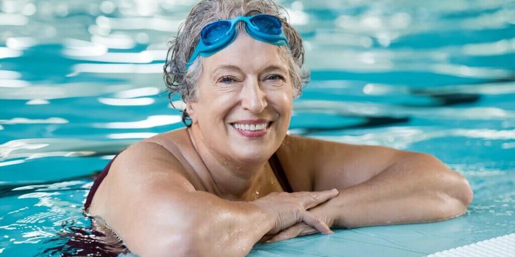 Mature woman wearing swim goggles at swimming pool. Fit active senior woman enjoying retirement standing in swimming pool and looking at camera. Happy senior healthy old woman enjoying active lifestyle.
